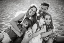 Erwachsene liebende Mann und Frau mit Sohn und Töchtern sitzen zusammen am Strand im Gegenlicht lächelnd in die Kamera — Stockfoto
