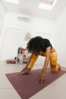 African American woman performing yoga pose with head down and stretching on mat in light room — Stock Photo