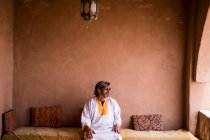 Uomo adulto in abiti lunghi seduto sul divano sulla terrazza con recinzione in pietra in stile orientale, Marocco — Foto stock