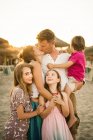 Adulto amante hombre y mujer besándose mientras sostiene un abrazo con el hijo y las hijas de pie juntos en la playa en la espalda iluminada - foto de stock
