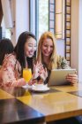 Jovem mulher caucasiana franzindo a testa e mostrando vídeo em tablet para surpreender amigo asiático enquanto sentados na mesa de café juntos — Fotografia de Stock