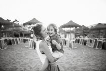 Vista lateral da mulher rindo carregando alegre filho brincalhão a mãos enquanto em pé na praia ao pôr do sol, foto em preto e branco — Fotografia de Stock