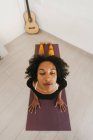 Cara de mujer joven afroamericana sentada en postura de yoga con los ojos cerrados en la estera en la sala de luz - foto de stock