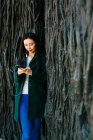 Прекрасна азіатка в стильному одязі, що слухає музику і користується смартфоном, спираючись на грубу стіну з рельєфом дерев. — стокове фото