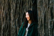 Femme asiatique excitée en tenue tendance souriante et regardant loin tout en s'appuyant sur le mur avec le relief des racines d'arbre — Photo de stock