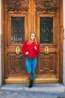 Positive junge Frau in stilvollem Outfit blickt in die Kamera, während sie sich an die Ziertür eines alten Gebäudes in der Stadtstraße lehnt — Stockfoto