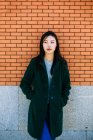 Pensive asiatique femme en tenue à la mode et regardant loin tout en s'appuyant sur le mur de briques — Photo de stock