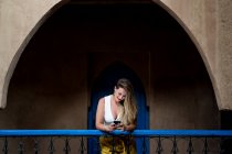 Alegre hermosa hembra en traje elegante sonriendo y navegando por el teléfono inteligente mientras se apoya en la barandilla del balcón del edificio antiguo en Marruecos - foto de stock