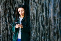 Hermosa mujer asiática con un atuendo elegante escuchando música y usando un teléfono inteligente mientras se apoya en la pared áspera con relieve de las rutas de los árboles - foto de stock