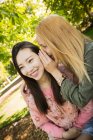Young Caucasian woman smiling and whispering secret into ear of smiling Asian friend while spending time in park together — Stock Photo
