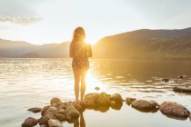 Вид ззаду на невідому жінку, що стоїть на скелі у воді, дивлячись на гори під час заходу сонця — стокове фото