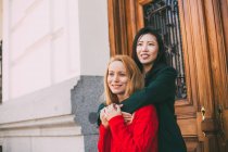 Glückliche asiatische Frau lächelt und schaut weg, während sie vor der Ziertür eines alten Gebäudes steht und ihren kaukasischen Freund umarmt — Stockfoto