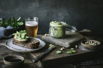Toasts with green cashew pate, herbs and slices of cucumber with jar and glass of beer on wooden board — Stock Photo