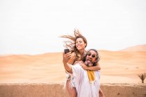 Мужчина средних лет с женщиной на спине делает селфи экспрессивно на террасе против песчаной пустыни, Марокко — стоковое фото