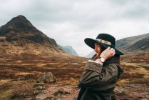 Hermosa mujer de jengibre usando y sosteniendo sombrero de pie contra pintorescas montañas de Escocia - foto de stock