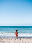 Back view of female kid in orange dress standing on sandy seaside and looking at sea and sky — Stock Photo