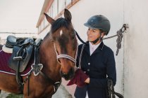 Side view of young teen woman in jockey helmet and jacket caressing horse standing together outdoors — Stock Photo