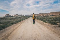 Seitenansicht eines jungen Mannes in gelber Jacke und Rucksack, der auf einer leeren Straße steht, die sich hoch zwischen steinigen Hügeln in Bardenas Reales Navarra erstreckt (automatische Übersetzung) — Stockfoto
