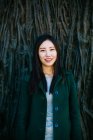 Emozionata donna asiatica in abito alla moda sorridente e distogliendo lo sguardo mentre si appoggia al muro con rilievo delle radici degli alberi — Foto stock
