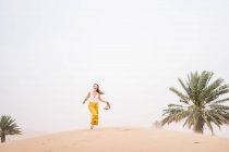 Cheerful stylish blonde woman holding shoes while walking in desert of Morocco — Stock Photo
