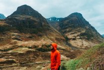 Side view of young man wearing a red raining coat hoodie while standing against picturesque mountains of Scotland — Stock Photo