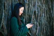 Beautiful Asian woman in stylish outfit listening to music and using smartphone while leaning on rough wall with relief of tree routs — Stock Photo