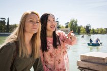 Multiethnic young women laughing and looking away while sitting on railing and pointing with finger near river — Stock Photo