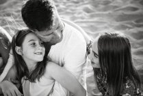 Hombre con hijas sentadas juntas en la playa con la espalda iluminada sonriéndose, foto en blanco y negro - foto de stock