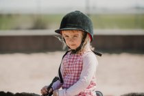 Menina pequena em vestido e jockey feno sentado no cavalo enquanto aprende a montar na pista de corrida — Fotografia de Stock