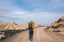 Visão traseira do jovem de jaqueta amarela e mochila andando em estrada vazia esticando alto entre colinas pedregosas em semi-deserto Bardenas Reales Navarra Espanha — Fotografia de Stock