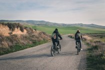 Back view of young men in dark clothes and backpack riding bicycles on empty road winding between stony hills in semi-desert Bardenas Reales Navarra Spain — Fotografia de Stock