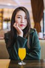 Азійка в стильному зеленому пальто дивиться на камеру, сидячи за столом у кафе зі склянкою свіжого соку. — стокове фото