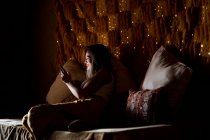 Seitenansicht einer jungen Frau, die in einem dunklen Raum in Marokko auf einem weichen Bett sitzt und lächelt und ihr Smartphone benutzt — Stockfoto