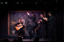 Homme en costume noir dansant le flamenco près des musiciens hispaniques masculins pendant la représentation contre la peinture sur scène sombre — Photo de stock
