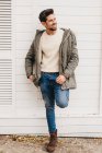 Trendy young man in jeans, boots and parka leaning on white wall and smiling outdoors — Stock Photo