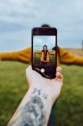 Imagen de la cosecha del hombre tatuado usando un teléfono inteligente para tomar una foto de una mujer joven con los brazos extendidos en el campo verde - foto de stock
