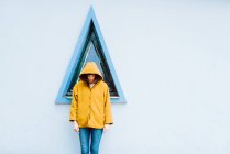 Jovem mulher de casaco quente amarelo sorrindo e olhando para baixo, enquanto está de pé contra a janela do triângulo e parede cinza do edifício — Fotografia de Stock