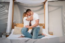 Вид збоку молодої романтичної пари в білих футболках і джинсах на колінах і обіймах в спальні гламуру — стокове фото