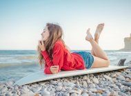 Seitenansicht der attraktiven lächelnden langhaarigen Frau in rotem Kapuzenpullover, die es genießt, auf einem Surfbrett am steinigen Meer zu liegen — Stockfoto