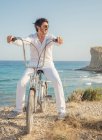 Ältere bärtige sportliche Mann mit Sonnenbrille beim Radfahren am Meer mit trockenem Gras vor dem Hintergrund der erstaunlichen türkisfarbenen Meereslandschaft in hellem Tag — Stockfoto