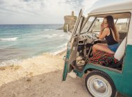 Вид сбоку на автомобиль с открытой дверью и женщину, рисующую на песчаном пустом пляже с пенными волнами в яркий солнечный день — стоковое фото