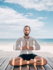 Adult bearded man meditating while sitting in lotus pose on wooden pier by seashore with legs crossed and looking at camera — Stock Photo