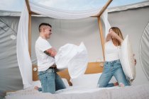 Alegre joven pareja divirtiéndose durante la pelea de almohadas en la cama en una tienda grande con techo transparente - foto de stock