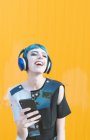 Mulher informal alegre em fones de ouvido e smartphone ouvindo música enquanto está de pé contra a parede amarela vívida — Fotografia de Stock