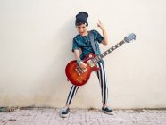 Дерзкий активный возбужденный мальчик в красочной одежде играет на гитаре, показывая два пальца на фоне белой стены — стоковое фото
