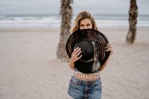 Cheerful blonde woman in colorful top and jean shorts blocking her face with a big hat on seashore — Stock Photo