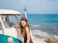 Mujer sonriendo y mirando hacia abajo cerca de blanco viejo coche a la orilla del mar en un día brillante y sereno - foto de stock
