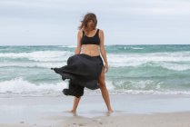 Attraktive Frau im schwarzen Outfit tanzt auf Sand in der Nähe des winkenden Meeres — Stockfoto