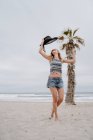 Attraktive Frau in Top und Shorts tanzt auf sandigem Meeresufer mit schwarzem Hut in erhobener Hand — Stockfoto