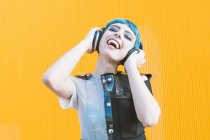 Joven alegre en vestido alternativo de moda sonriendo y escuchando música en auriculares contra la pared amarilla - foto de stock